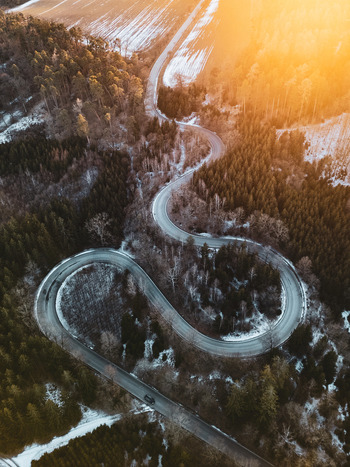 Aerial view of a serpentine road winding through a dense forest with patches of snow under the warm glow of the golden hour in Czechia.