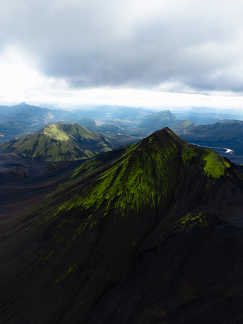 Aerial view of dark volcanic mountains rise sharply accented by patches of vibrant green moss under a cloudy sky Landmannalaugar Iceland. Aerial view of dark volcanic mountains rise sharply accented by patches of vibrant green moss under a cloudy sky Landmannalaugar Iceland.
