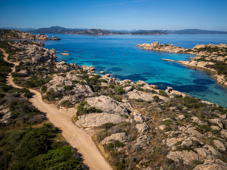 Aerial view of the rugged Carlotto coastline featuring turquoise waters granite rock formations and a winding dirt path under a clear blue sky in La Maddalena Sardinia Italy. Aerial view of the rugged Carlotto coastline featuring turquoise waters granite rock formations and a winding dirt path under a clear blue sky in La Maddalena Sardinia Italy.