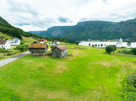 Aerial view of traditional wooden barns with grass roofs in a lush green meadow near a lake and a white church in the rural village of Araksbo Agder Norway.
