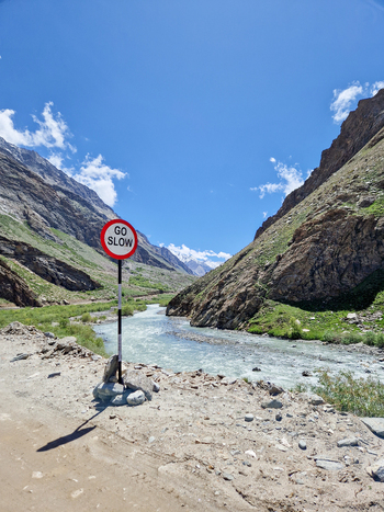 View of the Penzi La road featuring a GO SLOW sign beside a mountain river flowing through a rocky valley under a clear blue sky in Zanskar Ladakh India.