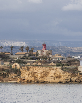 View of a red lighthouse standing on a rocky cliff with palm trees and coastal buildings under a cloudy sky in Syracuse Sicily Italy.