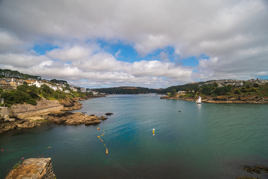 View of the Fowey River with sailing boats historic buildings and rocky coastlines under a cloudy sky in Fowey Cornwall United Kingdom. View of the Fowey River with sailing boats historic buildings and rocky coastlines under a cloudy sky in Fowey Cornwall United Kingdom.
