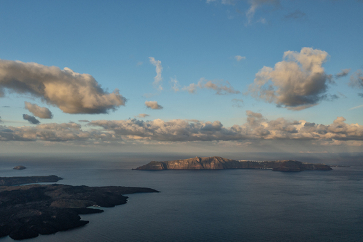 Aerial view of dramatic dark islands rising from tranquil deep blue waters under a vast sky painted with soft golden edged clouds Thira Greece. Aerial view of dramatic dark islands rising from tranquil deep blue waters under a vast sky painted with soft golden edged clouds Thira Greece.