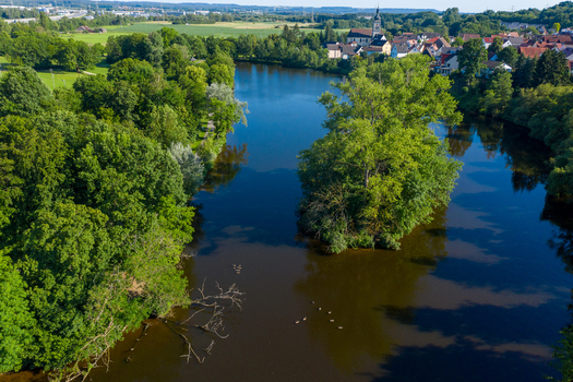 Aerial view of Sportinsel Forchheim Spazierweg und Spielplatz with a lush tree covered island in the river and a town skyline in Forchheim Bayern Germany.