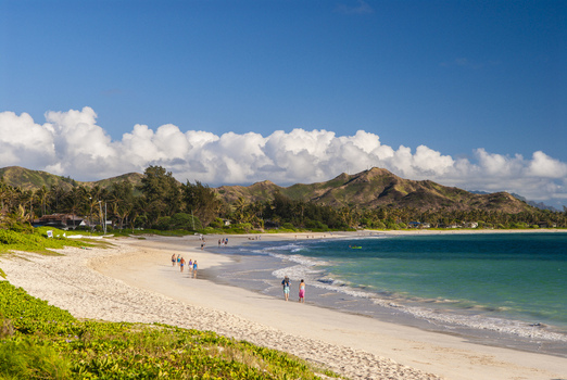 View of Kailua Beach with white sand turquoise water lush mountains and people walking along the shore under a bright blue sky in Kailua Hawaii United States.
