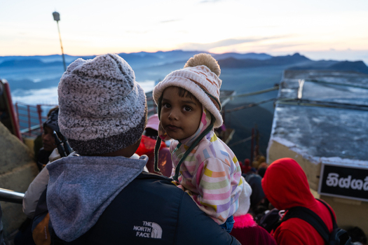 Ratnapura Sri Lanka 23 February 2026 View of a young child wearing a warm beanie being carried by an adult at the summit of Adam s Peak during sunrise with misty mountains.