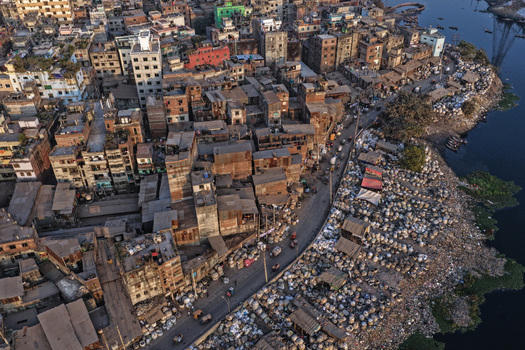 Aerial view of the Buriganga River flowing past the dense urban landscape of Dhaka featuring crowded buildings and a riverbank filled with goods in Dhaka Dhaka Division Bangladesh. Aerial view of the Buriganga River flowing past the dense urban landscape of Dhaka featuring crowded buildings and a riverbank filled with goods in Dhaka Dhaka Division Bangladesh.