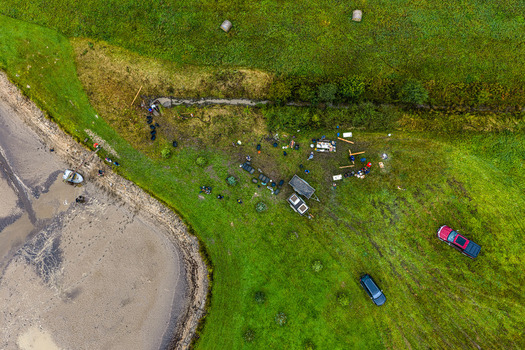 Latvia 26 September 2026 Aerial view of a group of people gathered in a grassy field near a shoreline with vehicles and equipment scattered across the landscape.