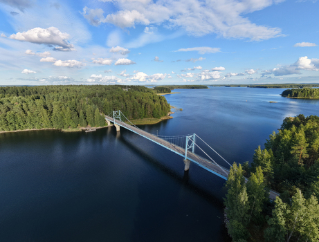 Aerial view of Potkusalmen silta Bridge spanning the calm waters of a lake surrounded by dense pine forests under a blue sky with clouds in Savonlinna Etela Savo Finland.
