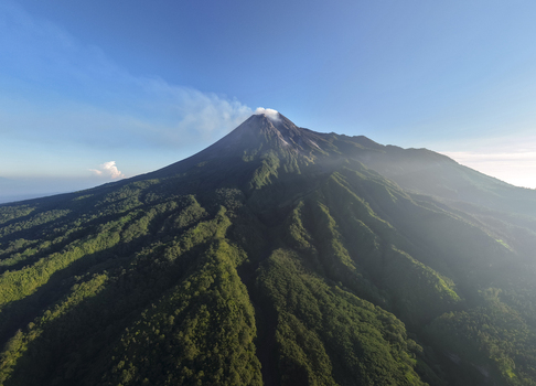 Aerial view of the Mount Merapi volcano with smoke rising from the crater above lush green forested slopes under a clear blue sky near Kecamatan Pakem Daerah Istimewa Yogya Indonesia. Aerial view of the Mount Merapi volcano with smoke rising from the crater above lush green forested slopes under a clear blue sky near Kecamatan Pakem Daerah Istimewa Yogya Indonesia.