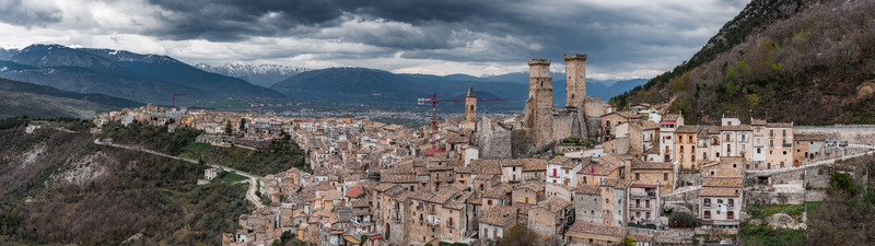 Aerial view of the medieval village of Pacentro featuring the Caldora Castle towers and historic stone houses under a dramatic cloudy sky with snow capped mountains in the distance Pacentro Abruzzo Italy. Aerial view of the medieval village of Pacentro featuring the Caldora Castle towers and historic stone houses under a dramatic cloudy sky with snow capped mountains in the distance Pacentro Abruzzo Italy.
