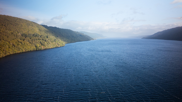 Aerial view of Loch Ness with deep blue waters rolling green hills and a soft cloudy sky in Highland Council Scotland United Kingdom.