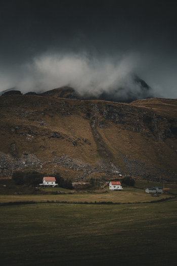 View of small houses with red roofs nestled at the base of a dramatic cloud covered mountain in the rural landscape of Leknes Nordland Norway. View of small houses with red roofs nestled at the base of a dramatic cloud covered mountain in the rural landscape of Leknes Nordland Norway.