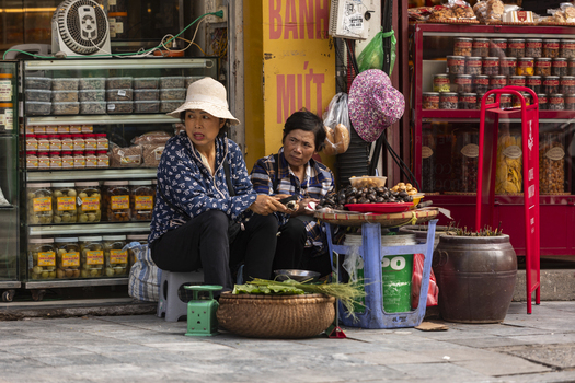 Hanoi Vietnam 11 November 2025 View of two Vietnamese women sitting on small plastic stools at a street market stall in Hanoi selling preserved fruits and snacks from glass jars and baskets.