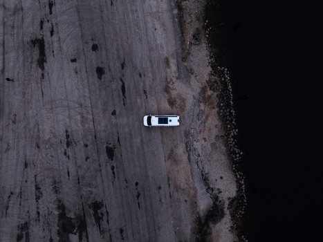 Aerial view of a white camper van parked on a gravel surface beside a dark lake in Latvia.