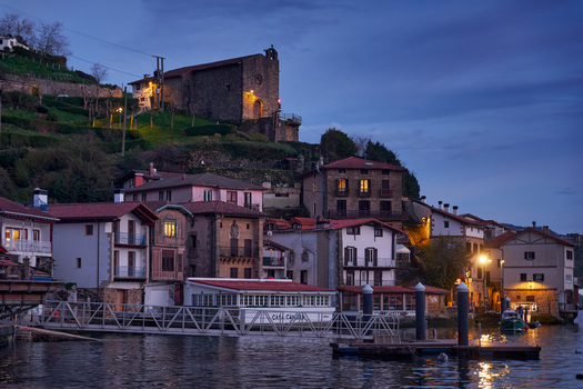 Pasai Donibane Spain 09 March 2026 View of the main harbor street along the coast with traditional buildings and the Ermita de Santa Ana church on the hill at dusk.