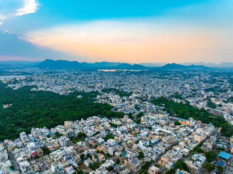 Aerial view of the city of Udaipur with dense urban buildings lush green parks and distant mountains under a soft sunset sky in Udaipur Rajasthan India. Aerial view of the city of Udaipur with dense urban buildings lush green parks and distant mountains under a soft sunset sky in Udaipur Rajasthan India.