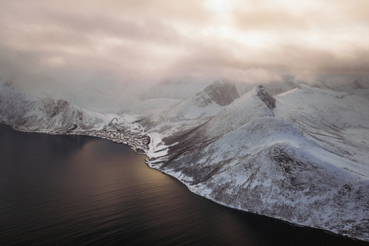 Aerial view of snow dusted mountain peaks meet the dark still waters under a clouded sky Fjordgård Troms Norway. Aerial view of snow dusted mountain peaks meet the dark still waters under a clouded sky Fjordgård Troms Norway.