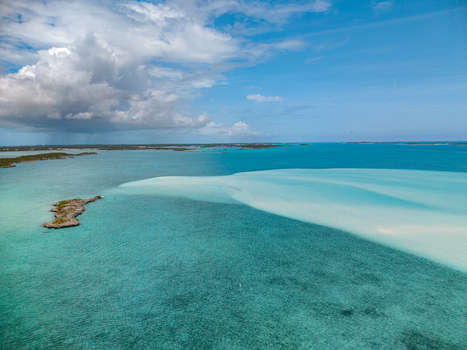 Aerial view of the Man O War Cay Sandbar stretching through turquoise waters near a small rocky island under a cloudy sky in Rolletown Exuma The Bahamas. Aerial view of the Man O War Cay Sandbar stretching through turquoise waters near a small rocky island under a cloudy sky in Rolletown Exuma The Bahamas.