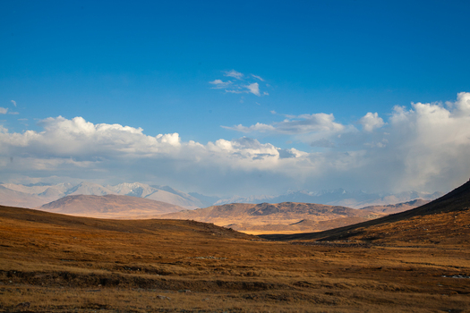 View of Deosai National Park with vast golden plains rolling hills and distant snow capped mountains under a bright blue sky with scattered clouds in Astore Gilgit Pakistan. View of Deosai National Park with vast golden plains rolling hills and distant snow capped mountains under a bright blue sky with scattered clouds in Astore Gilgit Pakistan.