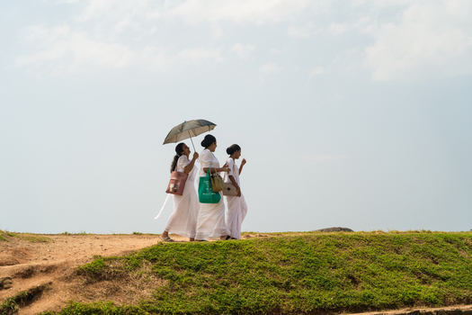 Sri Lanka 04 February 2026 View of three women dressed in traditional white clothing walking along a grassy ridge under a bright sky with one woman holding an umbrella.