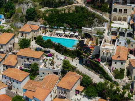 Aerial view of a luxury resort featuring a turquoise swimming pool and traditional stone buildings with terracotta rooftops on a lush hillside in Dhermi Vlore County Albania. Aerial view of a luxury resort featuring a turquoise swimming pool and traditional stone buildings with terracotta rooftops on a lush hillside in Dhermi Vlore County Albania.
