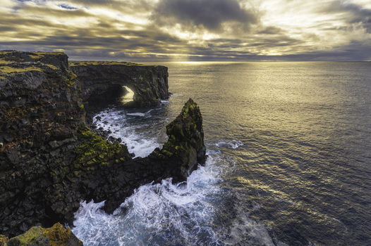 View of the rugged Svortuloft sea cliffs and natural rock arch with crashing waves under a golden sunset sky in Svortuloft Snaefellsbaer Iceland.