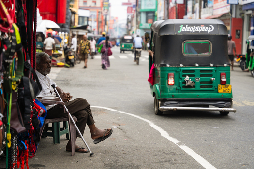 Sri Lanka 15 February 2026 View of an elderly man sitting with a walking cane on a busy street while a green auto rickshaw drives through a bustling urban market.