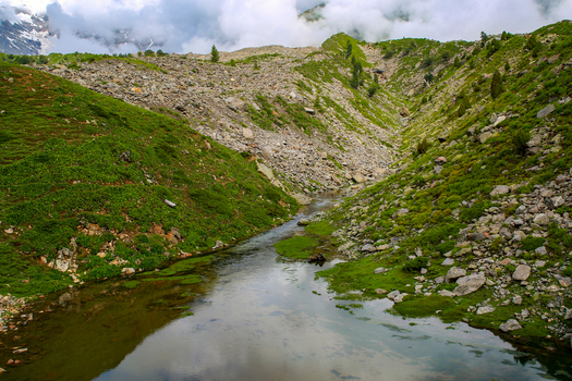 View of a stream flowing through the lush green landscape of Rama Meadows with rocky slopes and distant mountains under a cloudy sky in Astore Gilgit Pakistan. View of a stream flowing through the lush green landscape of Rama Meadows with rocky slopes and distant mountains under a cloudy sky in Astore Gilgit Pakistan.