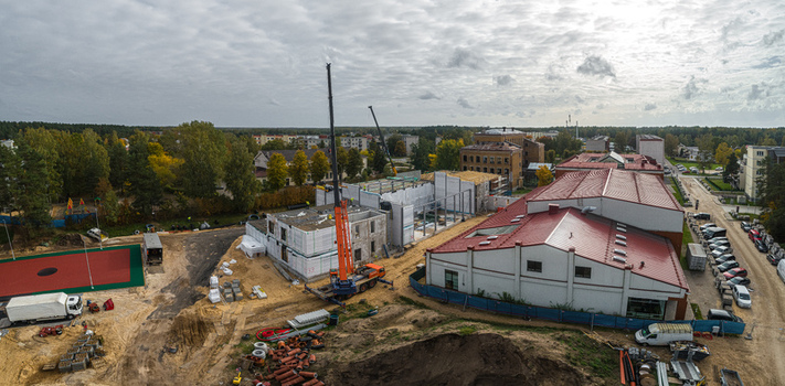 Aerial view of a construction site with a tall mobile crane unfinished building structures and a large facility with a red roof surrounded by trees and urban infrastructure in Latvia. Aerial view of a construction site with a tall mobile crane unfinished building structures and a large facility with a red roof surrounded by trees and urban infrastructure in Latvia.
