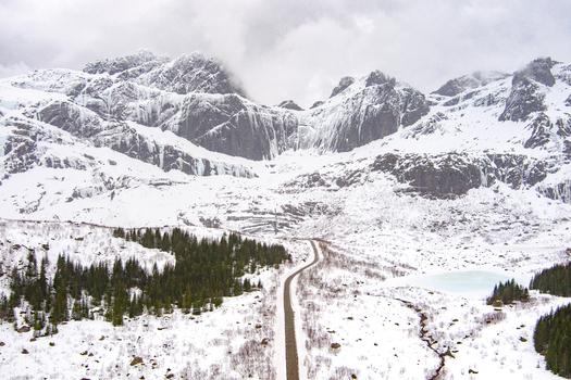 Aerial view of a winding road cutting through the snow covered landscape of Lofoten with coniferous trees and dramatic mountains in the background in Nordland Norway.
