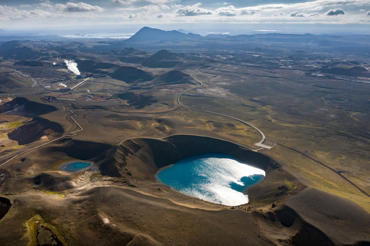 Aerial view of a deep blue volcanic crater lake surrounded by rugged dark terrain and a geothermal power plant in the distance Iceland.
