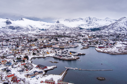 Aerial view of the snowy coastal town of Lofoten with colorful houses a harbor and dramatic snow capped mountains under a cloudy sky in Nordland Norway.
