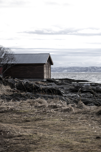 View of a traditional wooden boathouse on a rocky shore with snow capped mountains across the fjord under an overcast sky in Tromso Troms Norway.