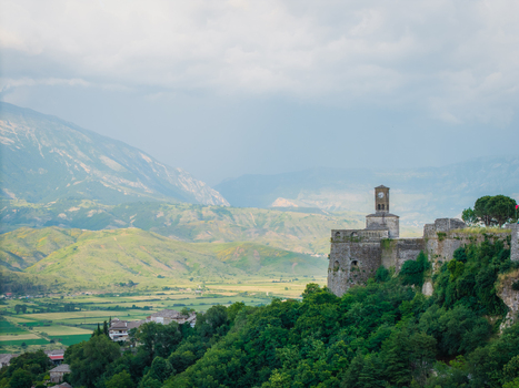 Aerial view of the historic Gjirokaster Castle featuring its iconic clock tower and stone fortifications overlooking a lush valley and mountains in Gjirokaster Gjirokaster County Albania. Aerial view of the historic Gjirokaster Castle featuring its iconic clock tower and stone fortifications overlooking a lush valley and mountains in Gjirokaster Gjirokaster County Albania.