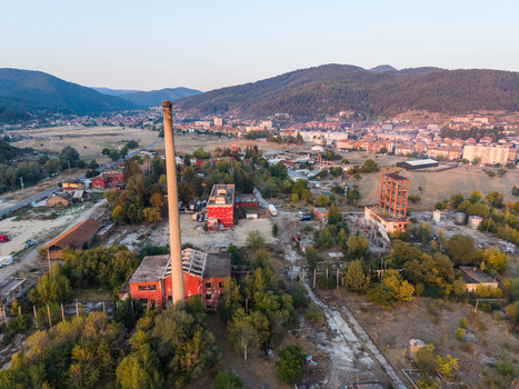 Aerial view of an abandoned industrial site featuring a tall chimney and red brick structures amidst overgrown vegetation with the town in the background Velingrad Pazardzhik Bulgaria. Aerial view of an abandoned industrial site featuring a tall chimney and red brick structures amidst overgrown vegetation with the town in the background Velingrad Pazardzhik Bulgaria.