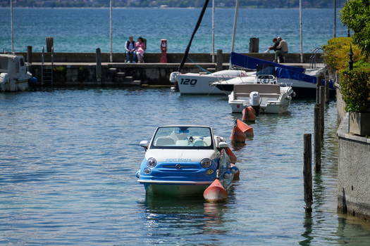 View of a unique boat shaped like a Fiat 500 car floating in the harbor with people sitting on the pier in Sirmione Lombardy Italy.