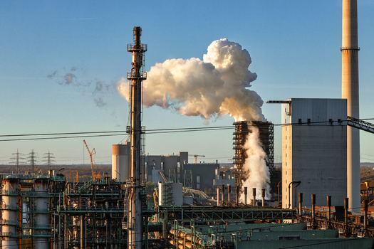 View of an industrial plant with thick white smoke rising from a chimney stack against a clear blue sky during sunset in Duisburg North Rhine Westphalia Germany.