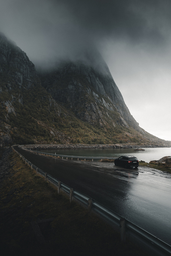 View of a black car parked on a wet coastal road beneath dramatic mist covered mountains in the Lofoten islands Nordland Norway. View of a black car parked on a wet coastal road beneath dramatic mist covered mountains in the Lofoten islands Nordland Norway.