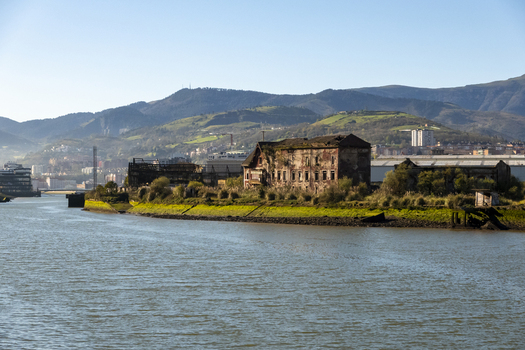 View of abandoned industrial buildings and ruins on the banks of the Nervion river with green hills under a clear blue sky Greater Bilbao Basque Country Spain. View of abandoned industrial buildings and ruins on the banks of the Nervion river with green hills under a clear blue sky Greater Bilbao Basque Country Spain.