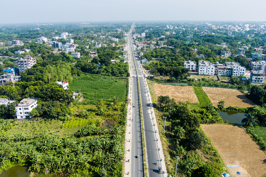 Aerial view of a long highway stretching through the urban landscape of Rajshahi surrounded by lush green fields scattered residential buildings and trees in Rajshahi Rajshahi Division Bangladesh.
