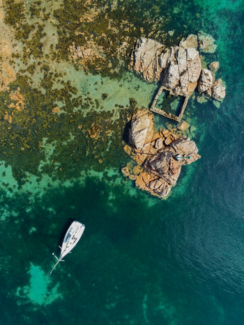 Aerial view of a white sailboat anchored in clear turquoise water near the rocky coastline and historic stone structures of Brehat Ile de Brehat Brittany France.