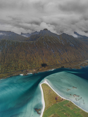 Aerial view of the dramatic Lofoten mountains rising above a turquoise bay with a sandy beach and lush green fields in Svolvaer Nordland Norway.