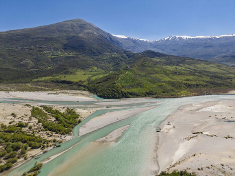 Aerial view of Vjosa Wild River National Park with turquoise braided river channels gravel banks green hills and snow capped mountains in Permet Gjirokaster County Albania. Aerial view of Vjosa Wild River National Park with turquoise braided river channels gravel banks green hills and snow capped mountains in Permet Gjirokaster County Albania.