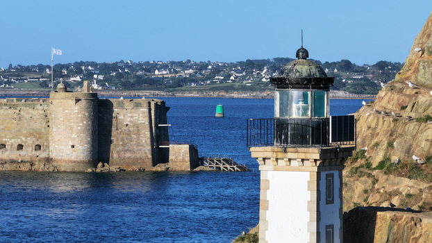 Aerial view of a white lighthouse on rugged cliffs overlooking the deep blue sea and a formidable stone fort Carantec Brittany France.