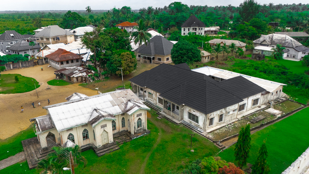 Aerial view of a residential area in Minima featuring a historic church surrounding houses with dark roofs lush green trees and people on sandy ground in Minima Rivers Nigeria.