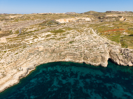 Aerial view of the rugged limestone cliffs and turquoise waters of Dwejra Bay with a winding coastal road and inland quarries under a clear blue sky in Gozo Region Malta. Aerial view of the rugged limestone cliffs and turquoise waters of Dwejra Bay with a winding coastal road and inland quarries under a clear blue sky in Gozo Region Malta.