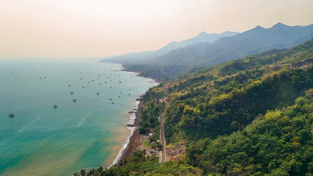 Aerial view of a winding coastal road along lush green hills a dark sand beach and numerous small fishing boats scattered across the turquoise sea in Sukabumi Indonesia. Aerial view of a winding coastal road along lush green hills a dark sand beach and numerous small fishing boats scattered across the turquoise sea in Sukabumi Indonesia.