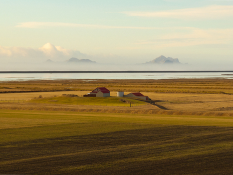 Aerial view of farm buildings with red roofs set in vast golden fields with distant mountains under a soft sky in Hvolsvollur Rangarthing eystra Iceland.